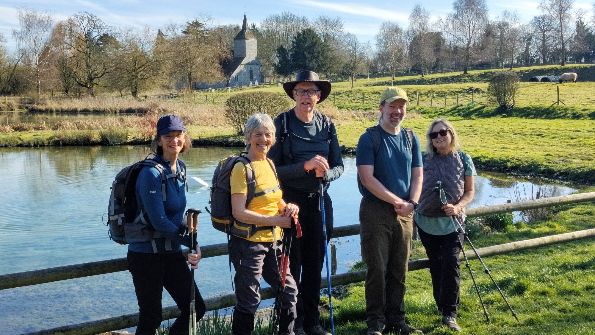 A group of 5 hikers standing in fromt of a pond. There is a small church and also trees in the background.